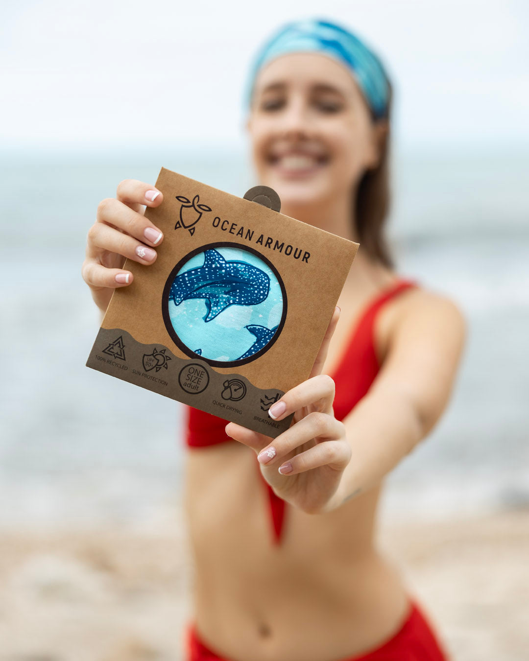 Person holding an Ocean Armour buff product with a whale shark design on it , standing on a beach.