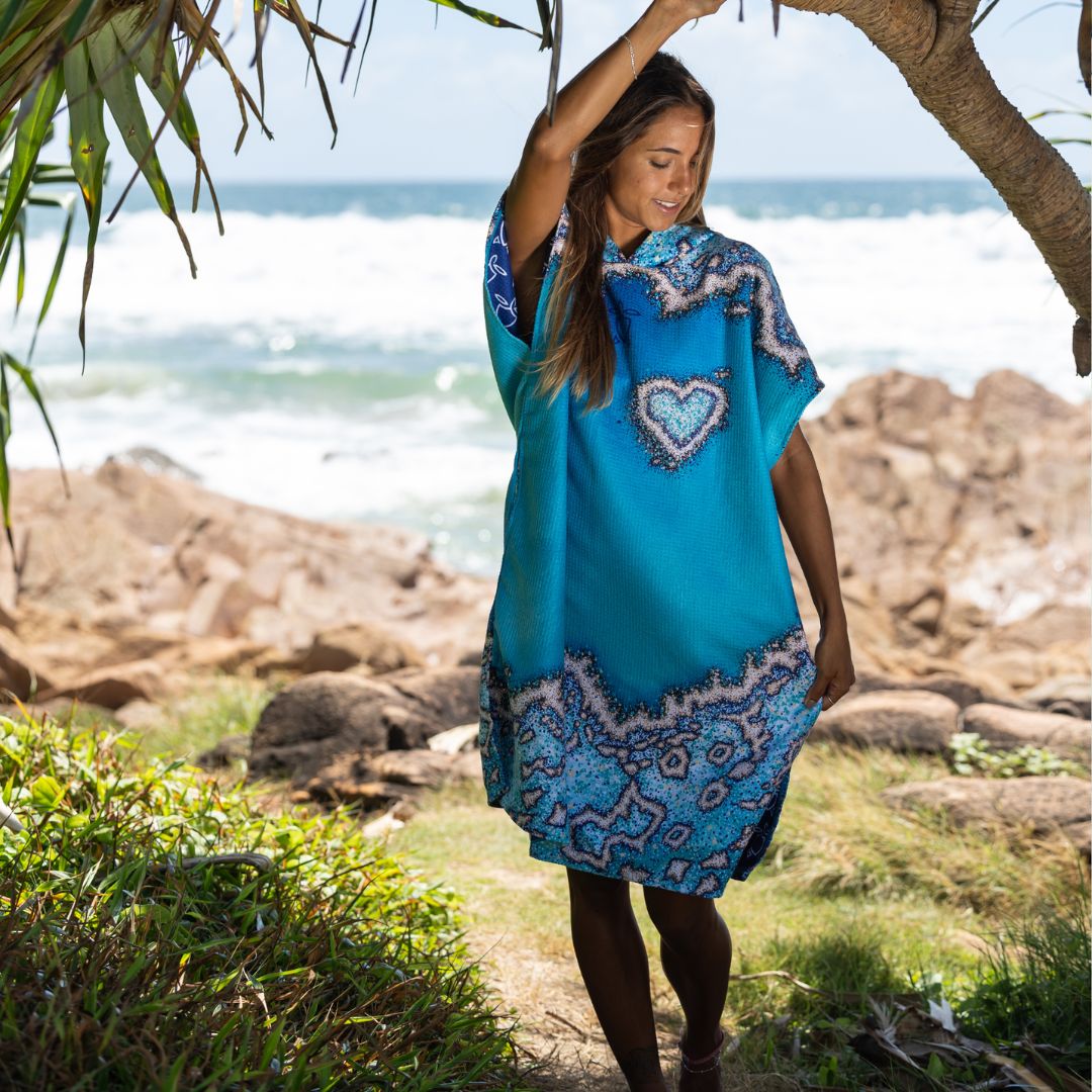 A woman wearing a bright turquoise blue kaftan dress with a heart-shaped chest motif and intricate paisley patterns, standing outdoors on a rocky beach path with the ocean and trees in the background.