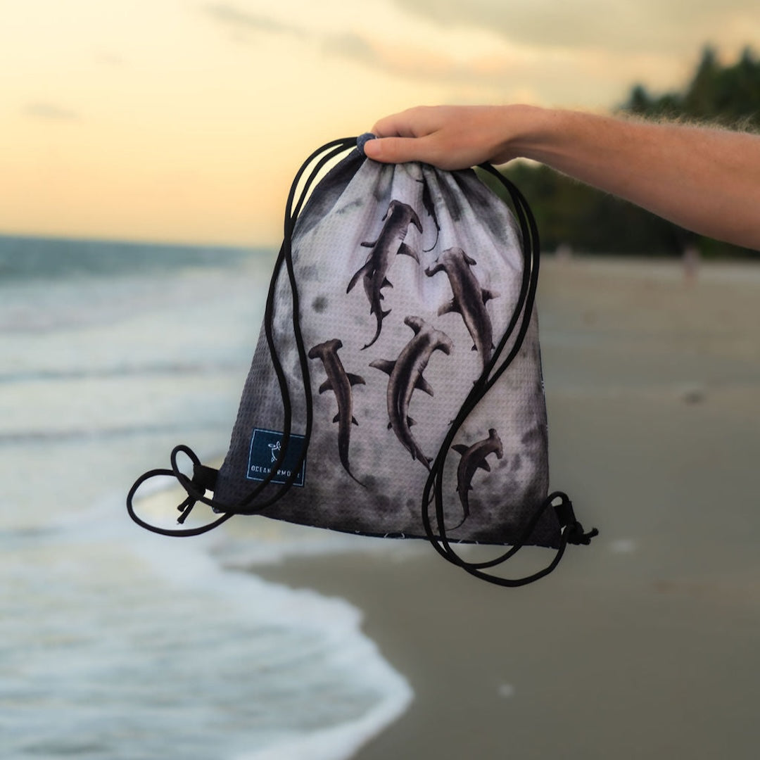 A drawstring gym bag with a shark print is held up by a person's hand on a beach at sunset. The bag has black drawstrings and a small blue brand tag that reads Shark Reef.