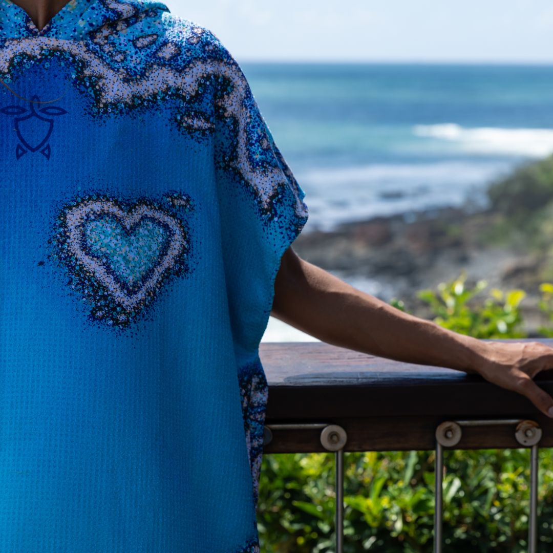A person wearing a bright blue ka'ole shirt with a heart and wave design, leaning on a railing with a scenic ocean background.