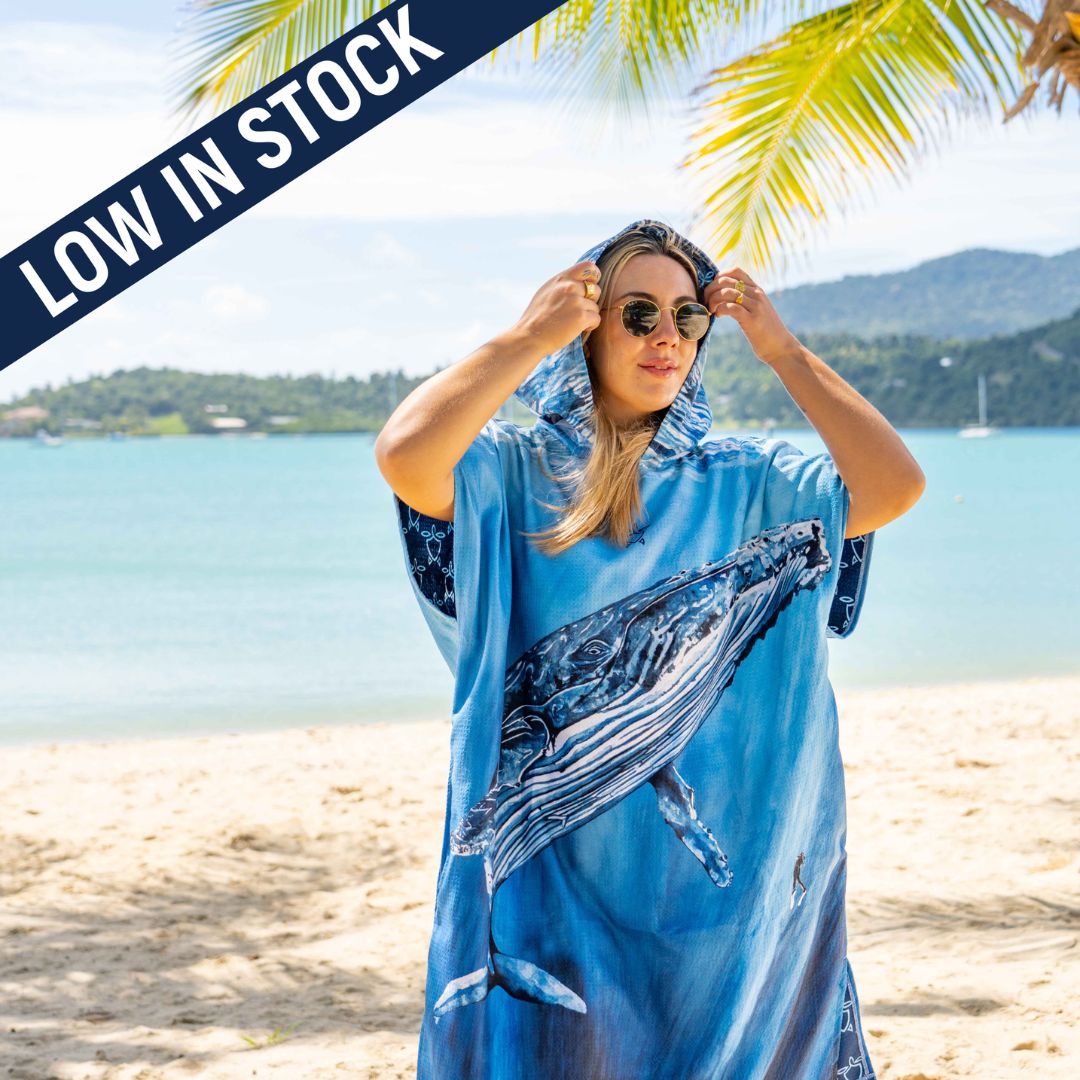 A woman stands on a sandy beach wearing a blue whale-print beach cover-up. The background shows the ocean, distant hills, and palm leaves.