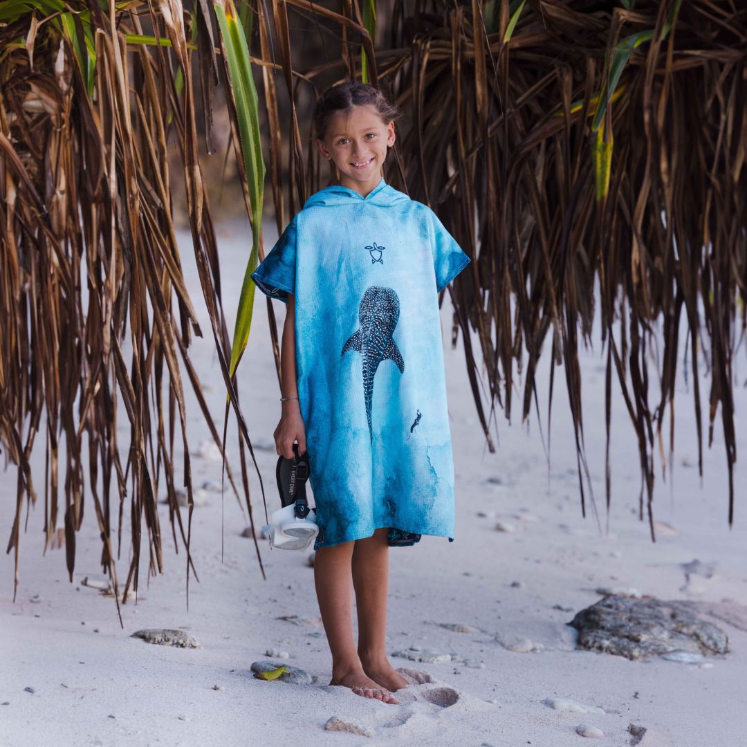 A young girl stands on a sandy beach under a palm thatch, wearing a blue hooded poncho-style cover-up with a whale shark graphic print.