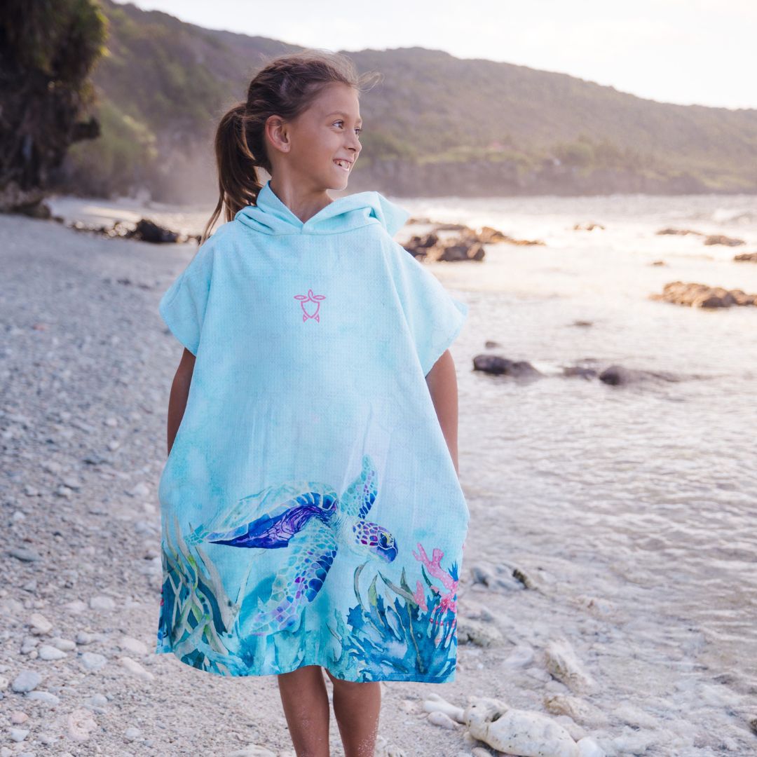 A young girl stands on a rocky beach wearing a light blue hooded poncho with a sea turtle and ocean wave print. The background shows the ocean and distant hills.