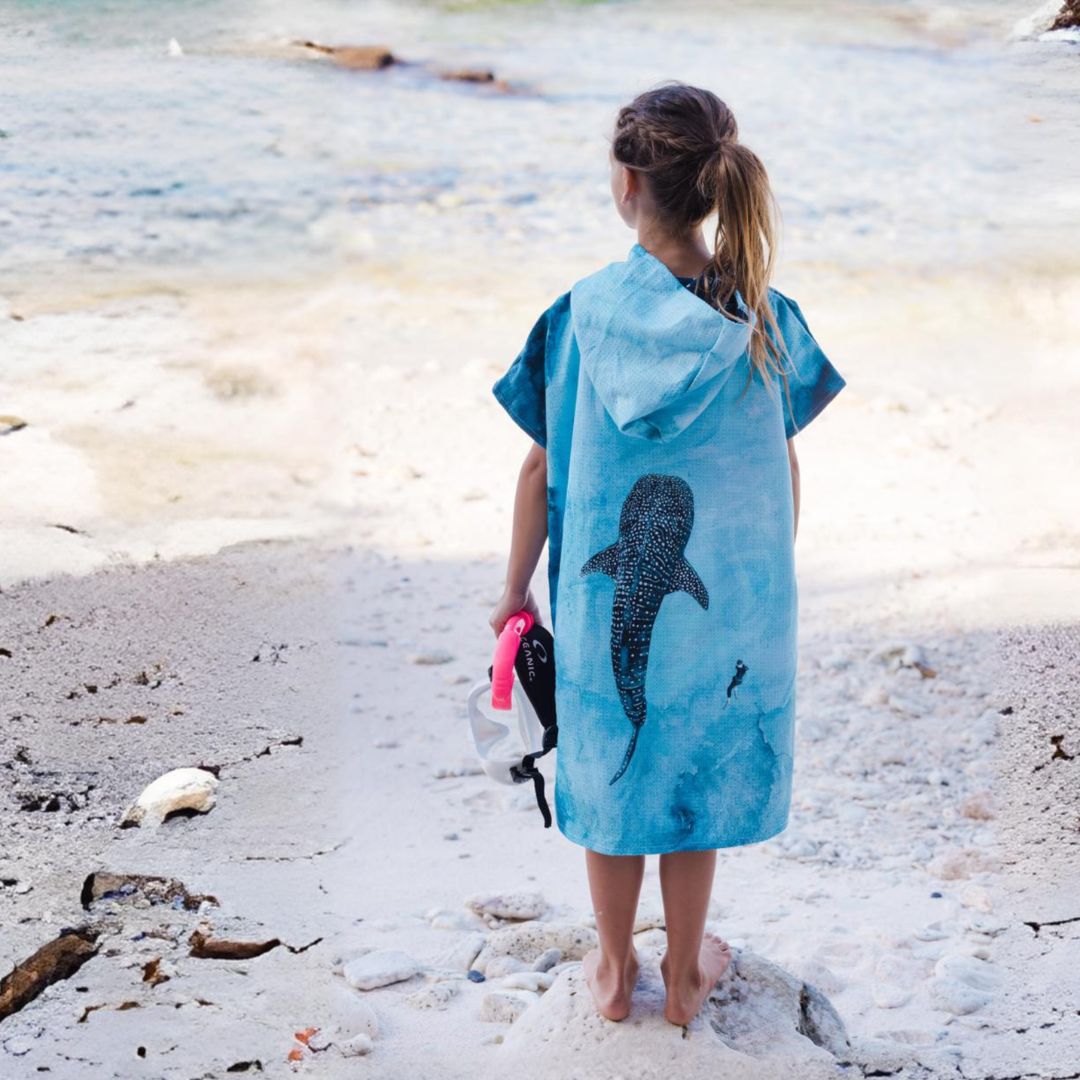 A child stands on a sandy beach facing the ocean, wearing a light blue hooded towel or poncho with a large whale shark print on the back. The child is holding a pink and black swim cap or goggles.
