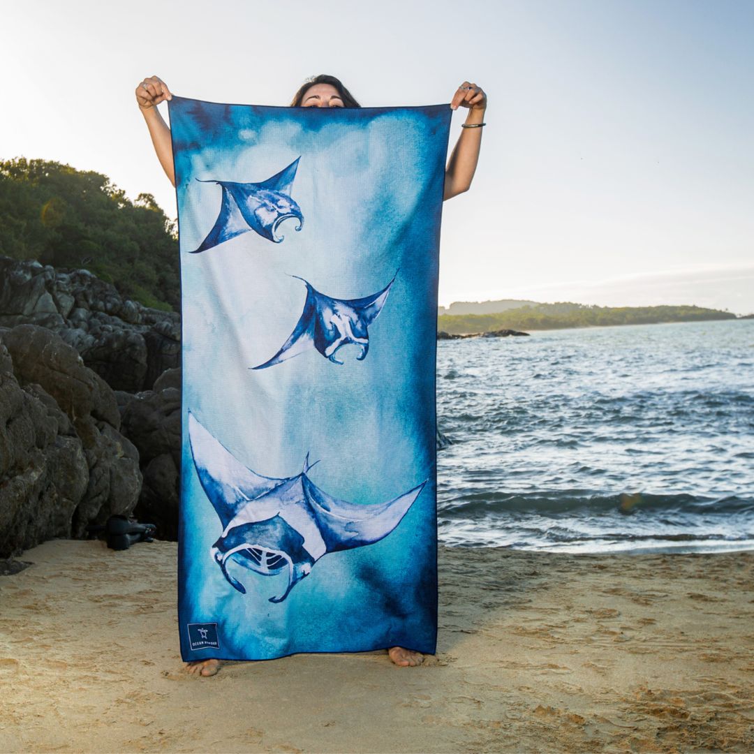 A woman stands on a sandy beach holding up a large blue beach towel printed with three manta rays swimming upwards against a water and shoreline background.