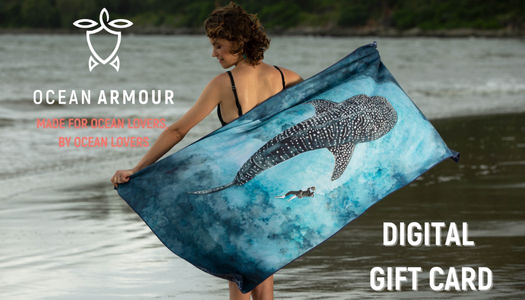 Female model holding a Whale Shark beach towel beside the ocean with Ocean Armour Digital Gift Card branding.