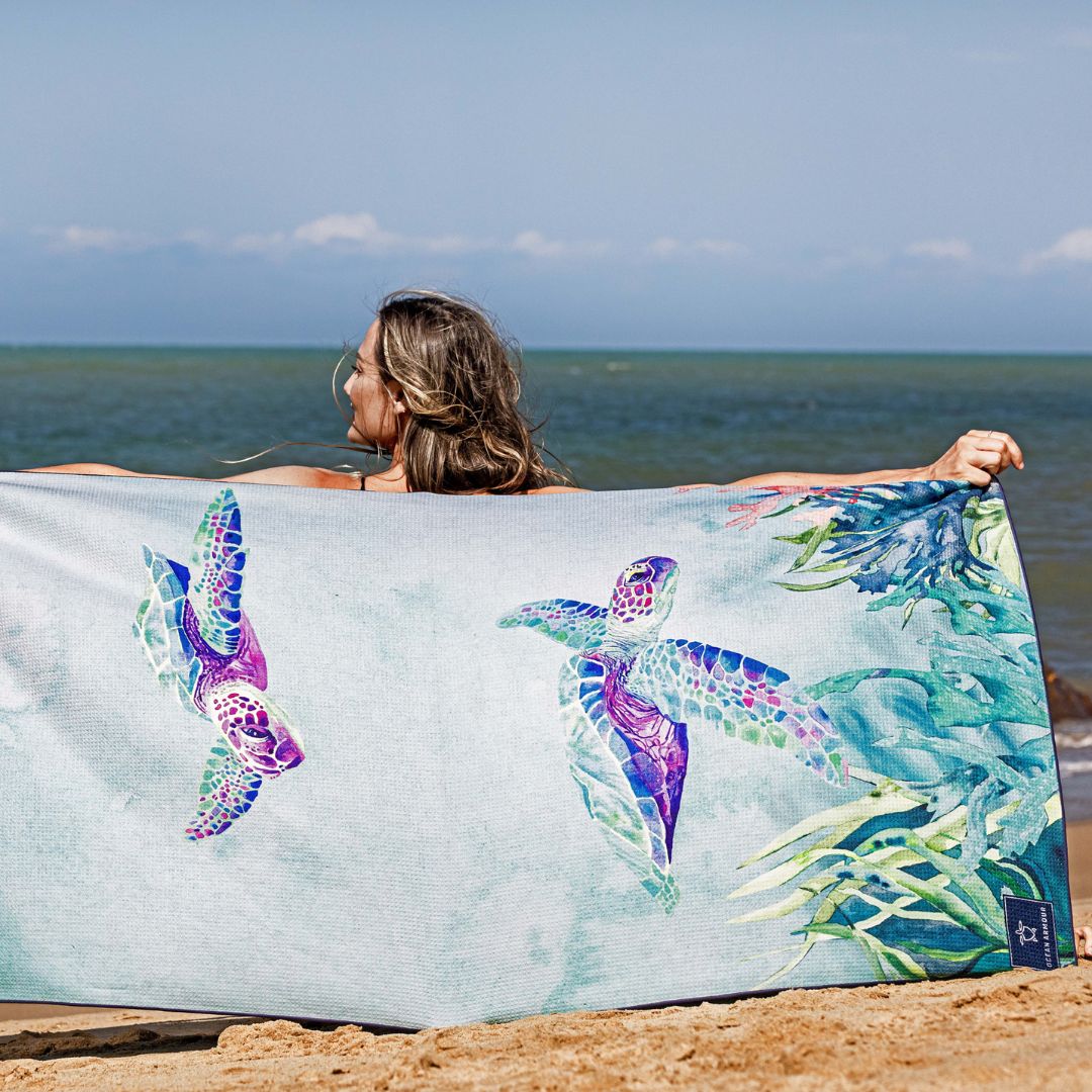A woman on a sandy beach holds up a large light blue beach towel featuring colorful sea turtle and tropical leaf illustrations.
