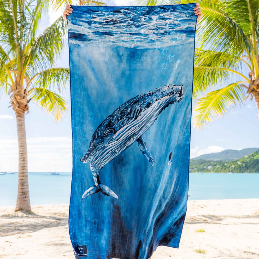 A blue beach towel featuring a printed whale underwater scene, held up by a person at the top corners against a tropical beach background with palm trees and ocean.
