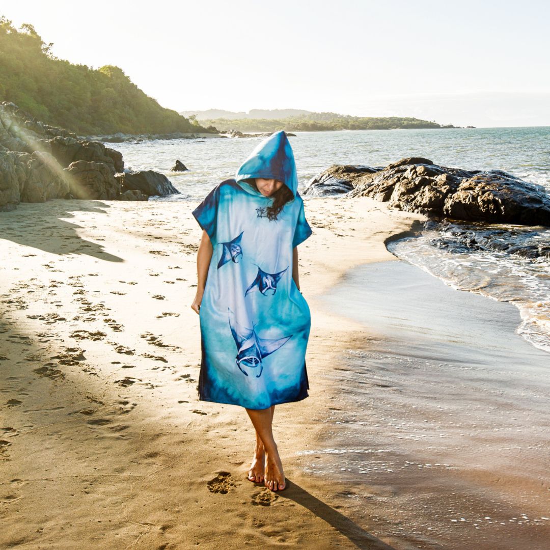 A woman stands on a sandy beach wearing a hooded blue and white printed poncho or cover-up with shark motifs, with ocean water, rocks, and shoreline in the background.