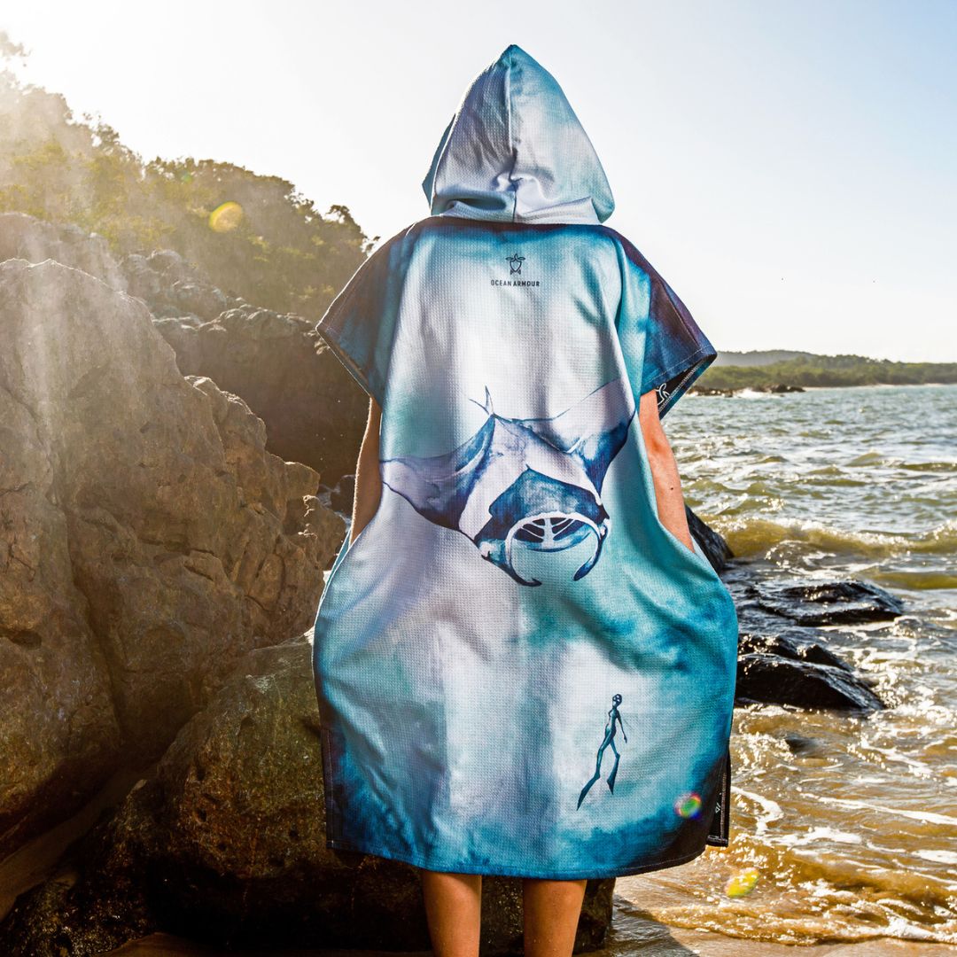 A person standing on a rocky beach with water and shoreline in the background, wearing a hooded poncho-style garment shown from the back. The garment has a blue, white, and gray printed design featuring a large fish motif and a small human silhouette near the lower right.