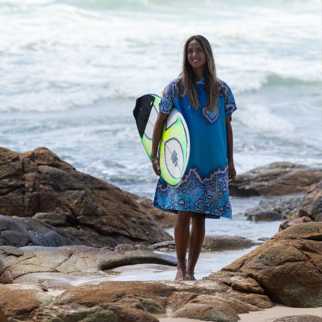 A woman stands on a rocky beach holding a surfboard, wearing a loose blue patterned dress or poncho-style garment with a heart motif on the chest.