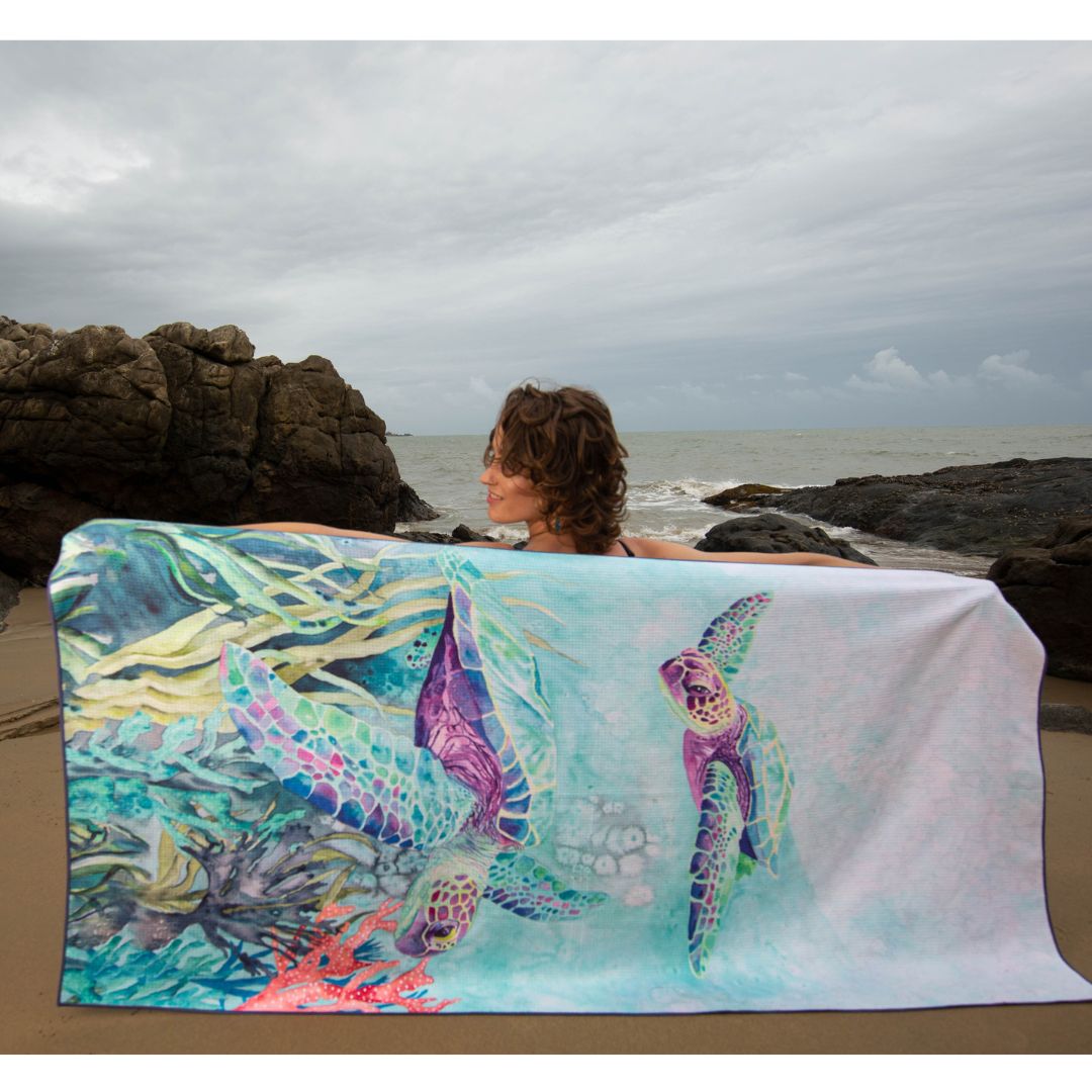 Ocean Armour Sea Turtle beach towel featuring detailed sea turtle artwork in shades of blue, green, and purple, displayed on a beach by a female model.