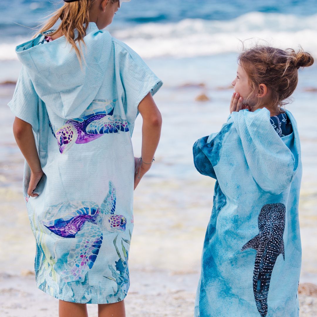 Two people standing on a beach, wrapped in matching light blue hooded towels featuring large sea turtle and whale shark prints.