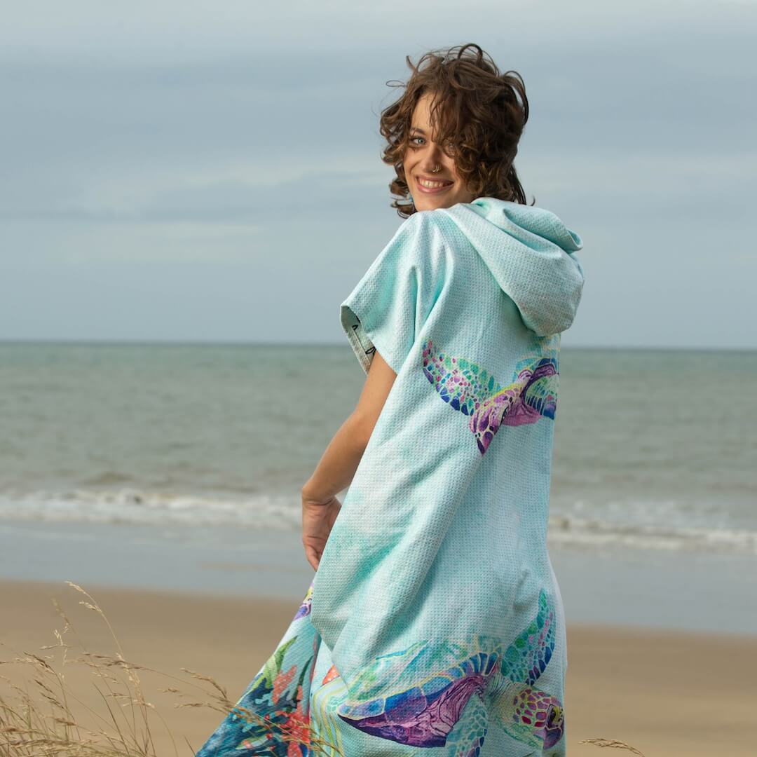 A woman standing on a sand dune wearing a light aqua hooded beach towel poncho with colorful turtle prints, looking back over her shoulder with a smile. The background shows the ocean.