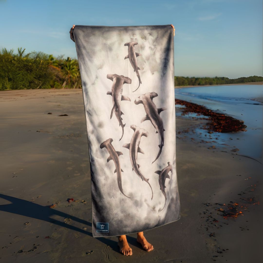 Person holding a Hammerhead Shark sand-free beach towel vertically on a sandy beach, featuring a watercolor-style hammerhead shark design on a gray ocean-inspired background.