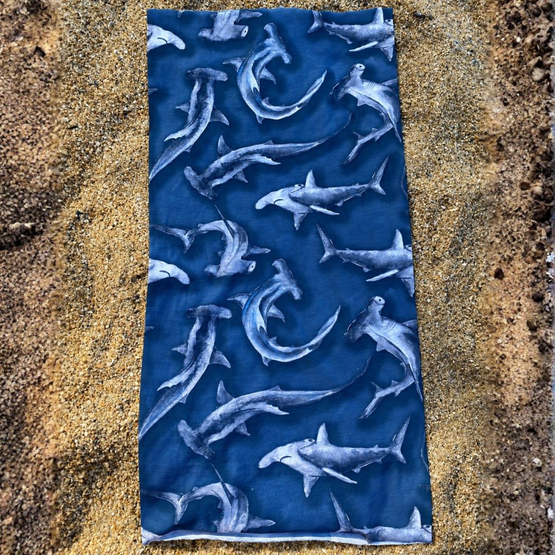 Hammerhead Shark Multi-Use Sun Buff with a navy blue background and an all-over hammerhead shark print, displayed flat on sand