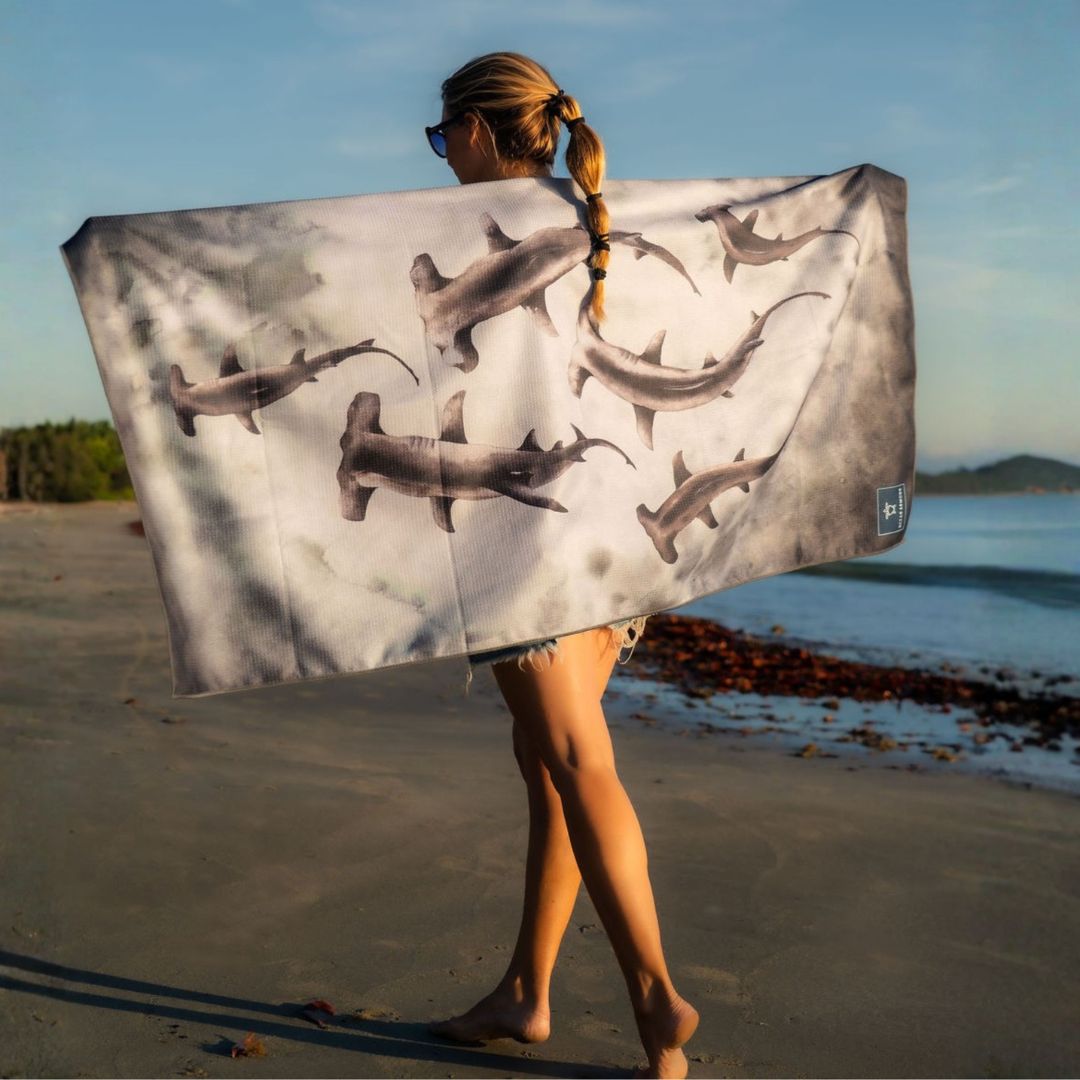 Hammerhead Shark beach towel with a watercolor-style shark pattern, held open outdoors on a beach with the ocean in the background.