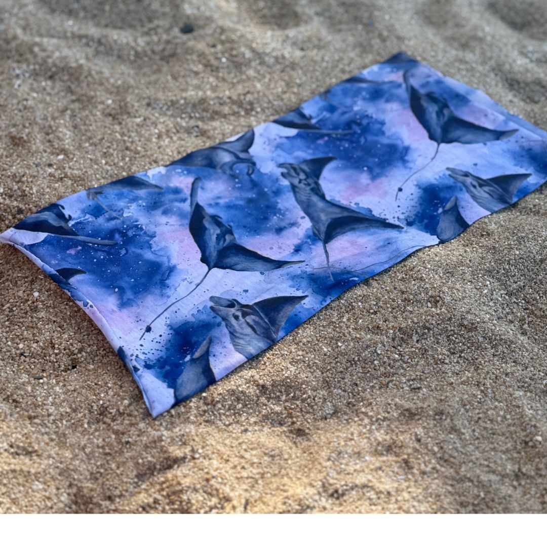 Blue multi-use Sun Buff featuring an all-over manta ray print in a watercolor-style ocean pattern, laid flat on sand