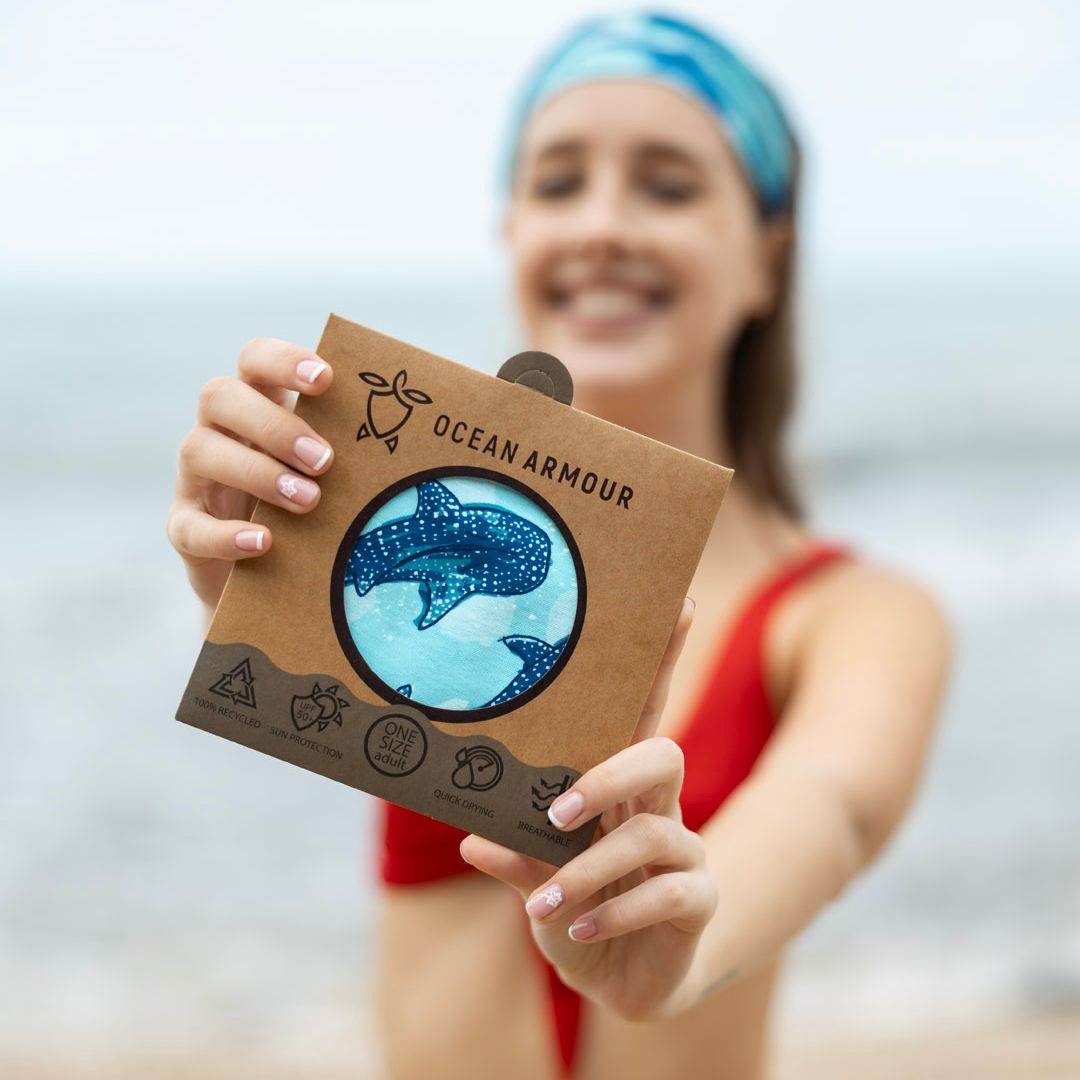 A woman on a beach holds up a brown cardboard package featuring a circular cutout with a blue spotted whale shark illustration. The package is branded Ocean Armour.