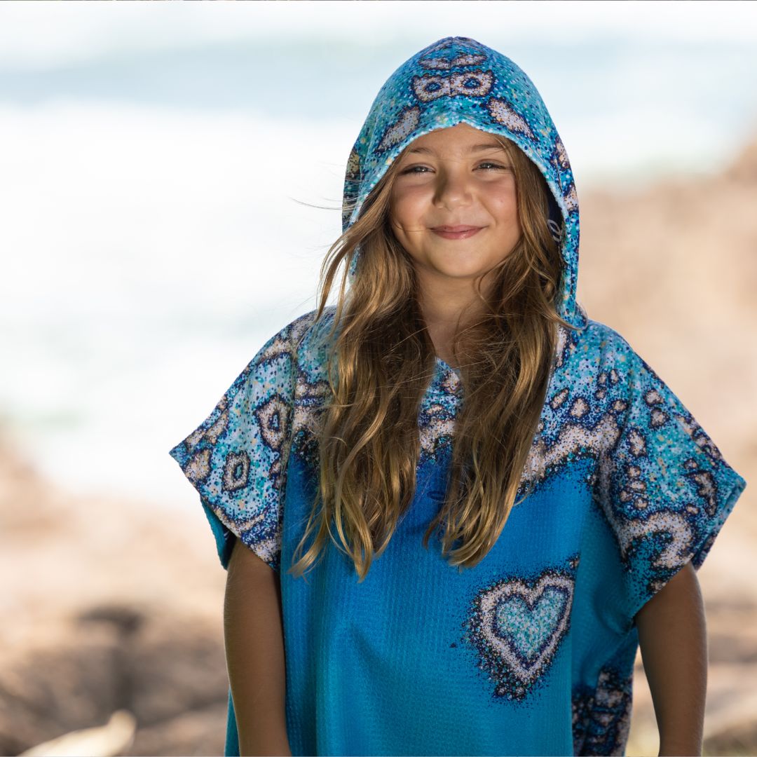 A young girl smiling while wearing a blue patterned hooded poncho top with a heart design, set against a blurred beach background.