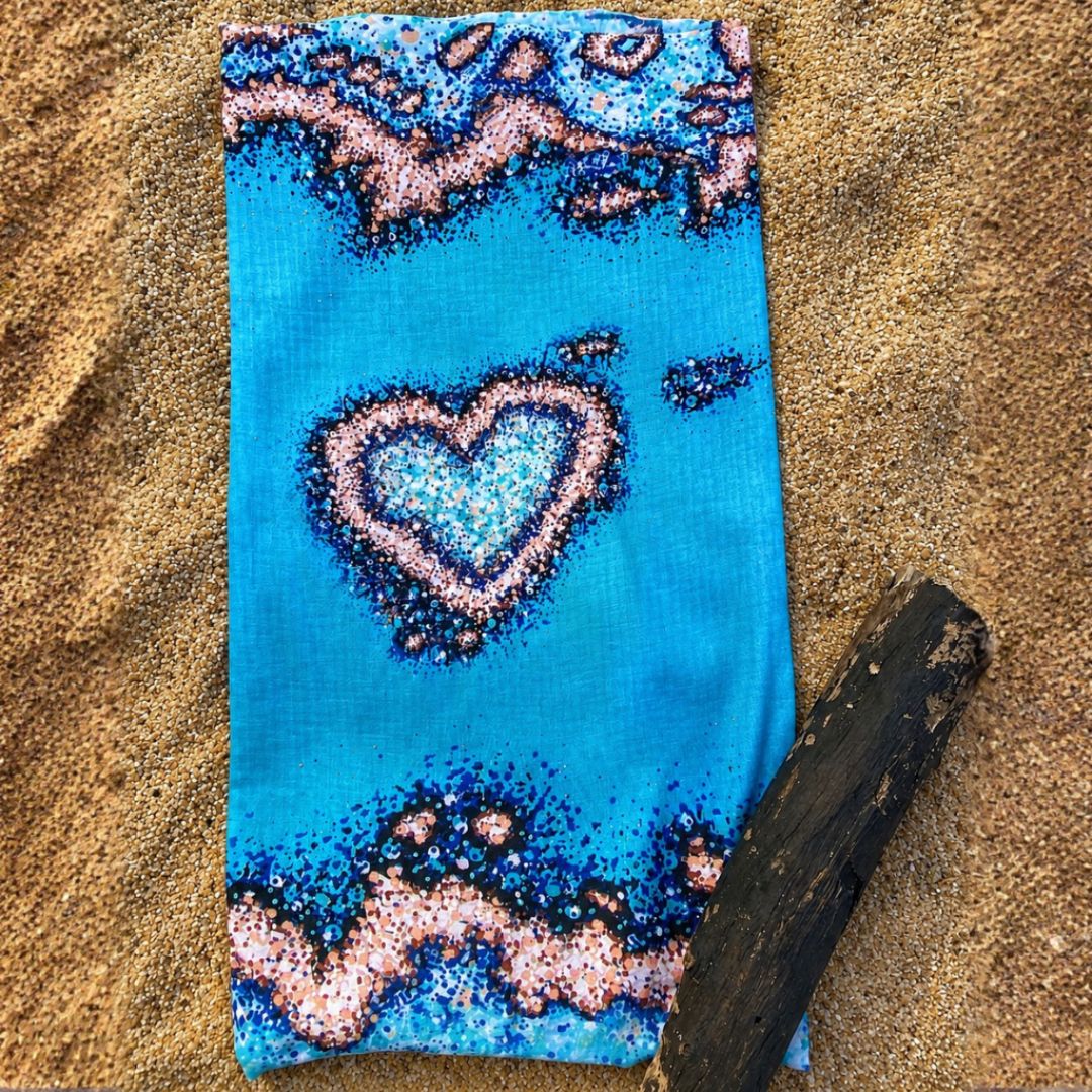 Multi-use Sun Buff with a blue ocean-inspired design and a heart-shaped coral reef pattern, displayed flat on a sandy beach