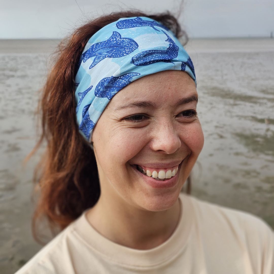 A smiling woman wearing a light blue and white patterned swim headband with dark blue shark designs, photographed outdoors against a blurred beach or water background.