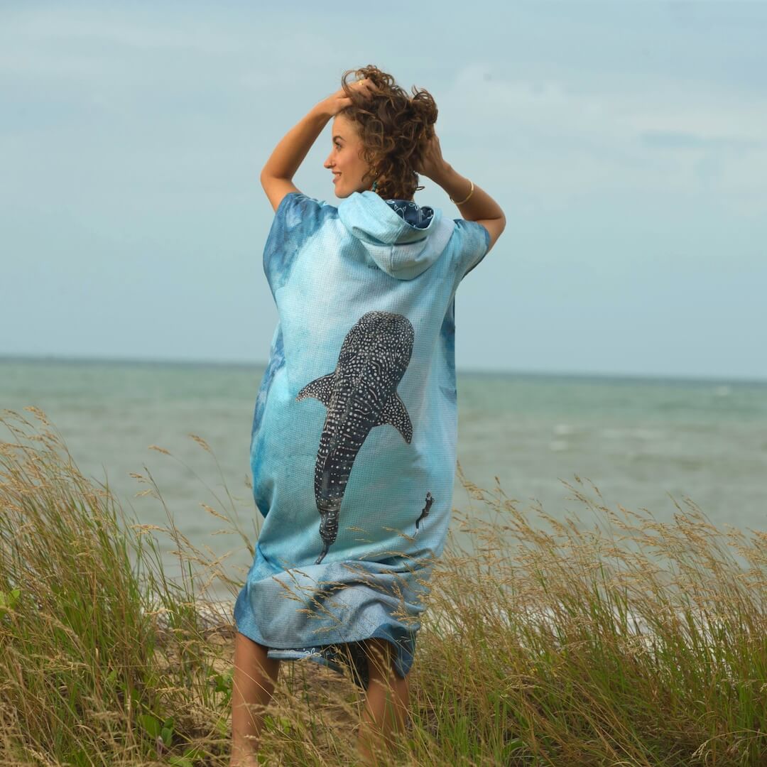 A woman stands on a beach in tall grass, wearing a light blue hooded towel poncho with a large watercolour whaleshark graphic on the back.