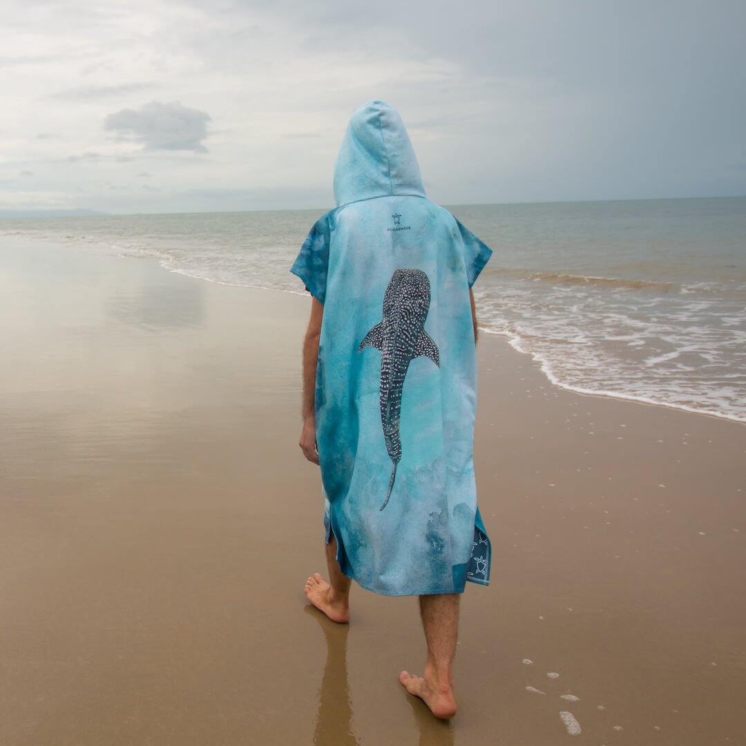 A person standing on a sandy beach facing the ocean, wearing a hooded blue towel poncho with a large whale shark watercolour design printed on the back.