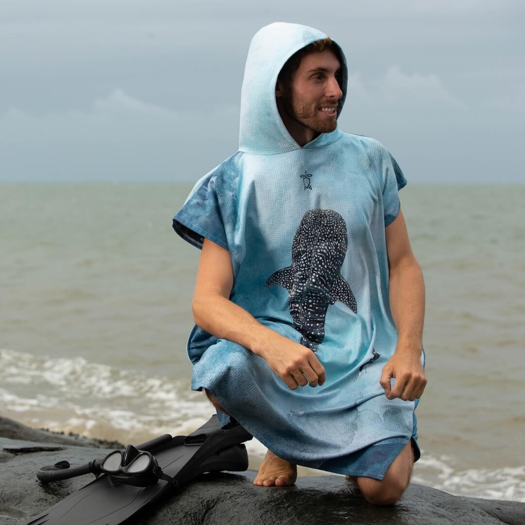 A man wearing a light blue hooded towel poncho with a whaleshark graphic sits on a rock at the beach, with snorkelling fins and snorkel beside him.