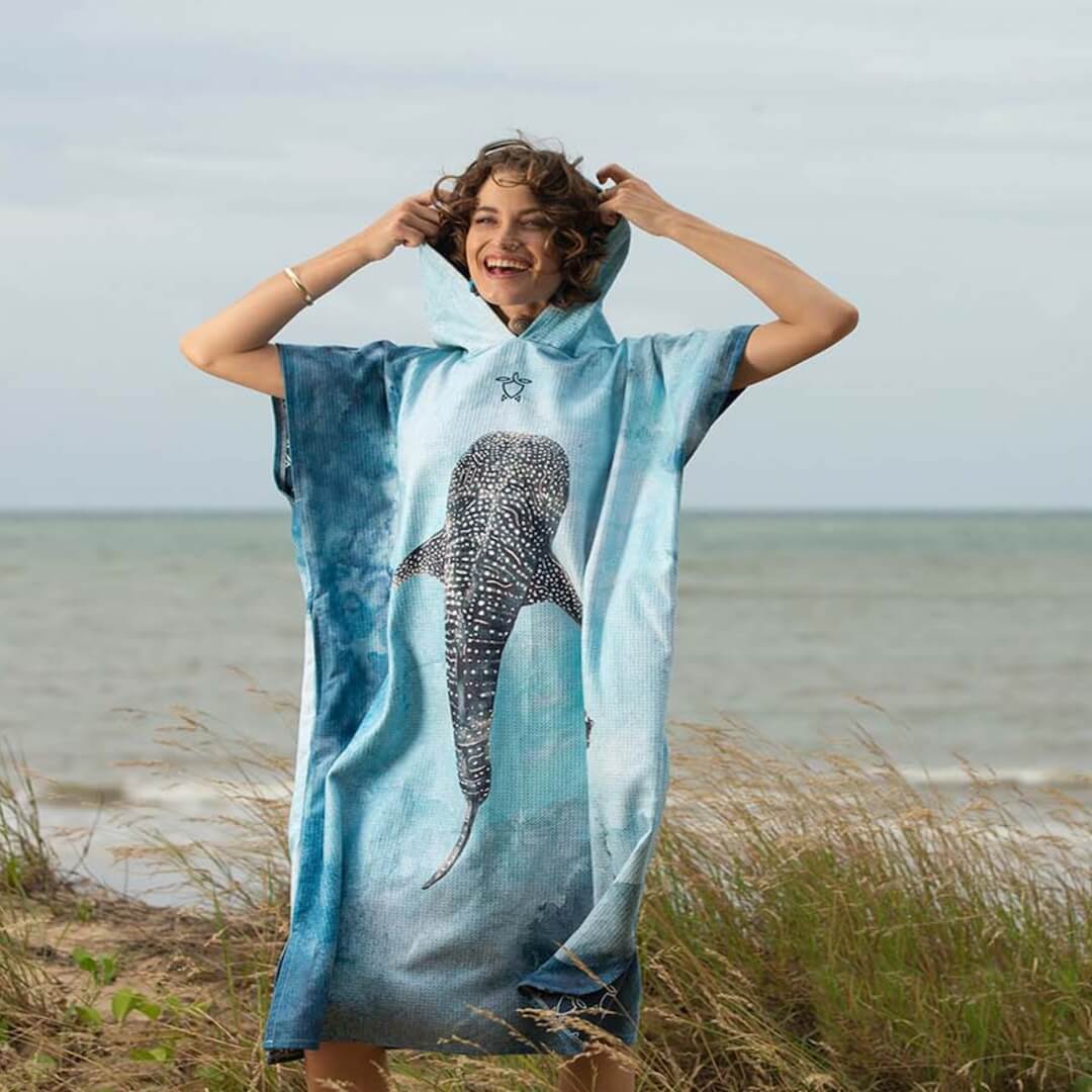 A woman stands on a sand dune wearing a blue hooded towel poncho with a large whale shark design on the front. The background shows the ocean and sky.