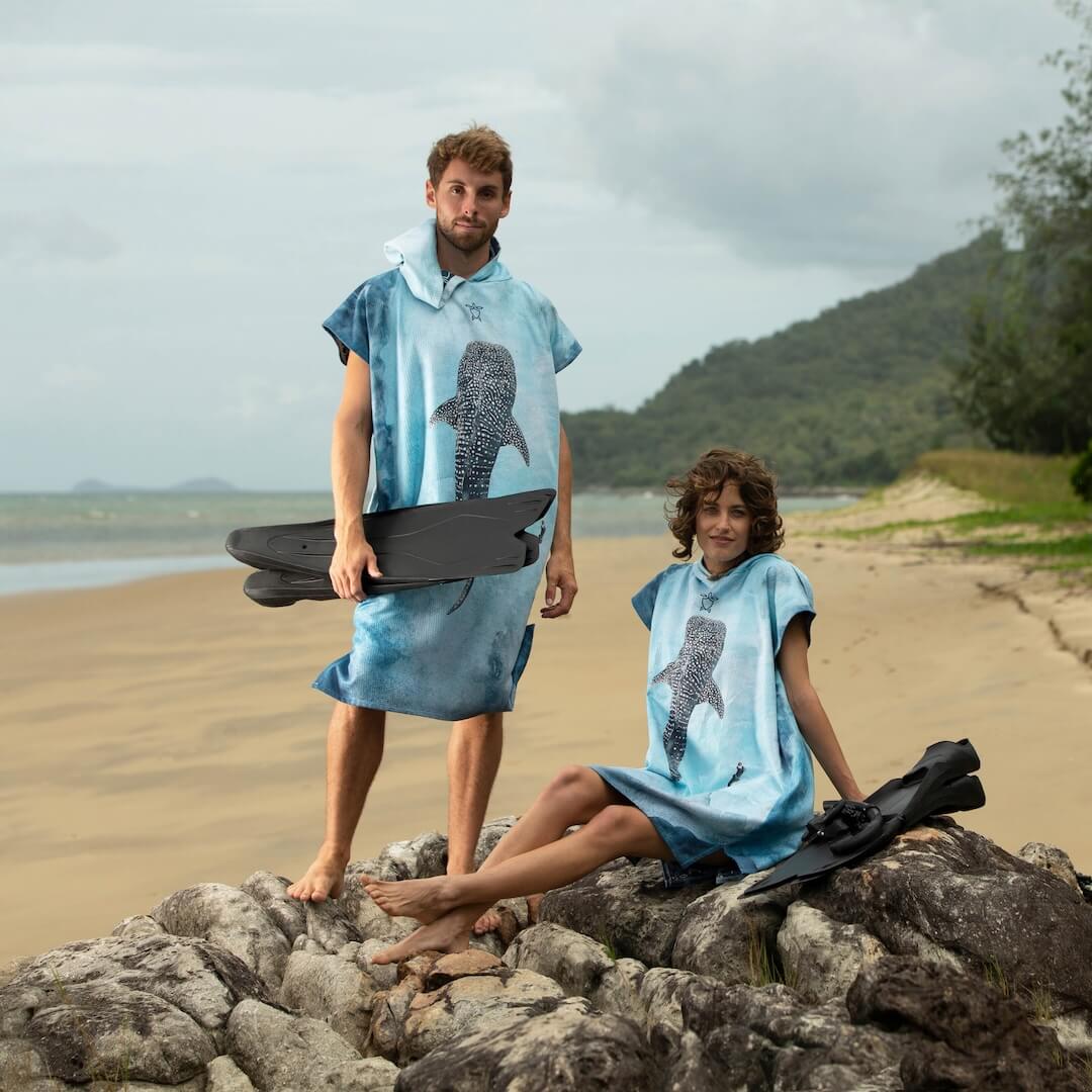 A man and a woman wearing matching light blue towel ponchos with a whaleshark design, standing on rocks at a beach. The man holds black snorkelling fins, and the woman also has black fins beside them.
